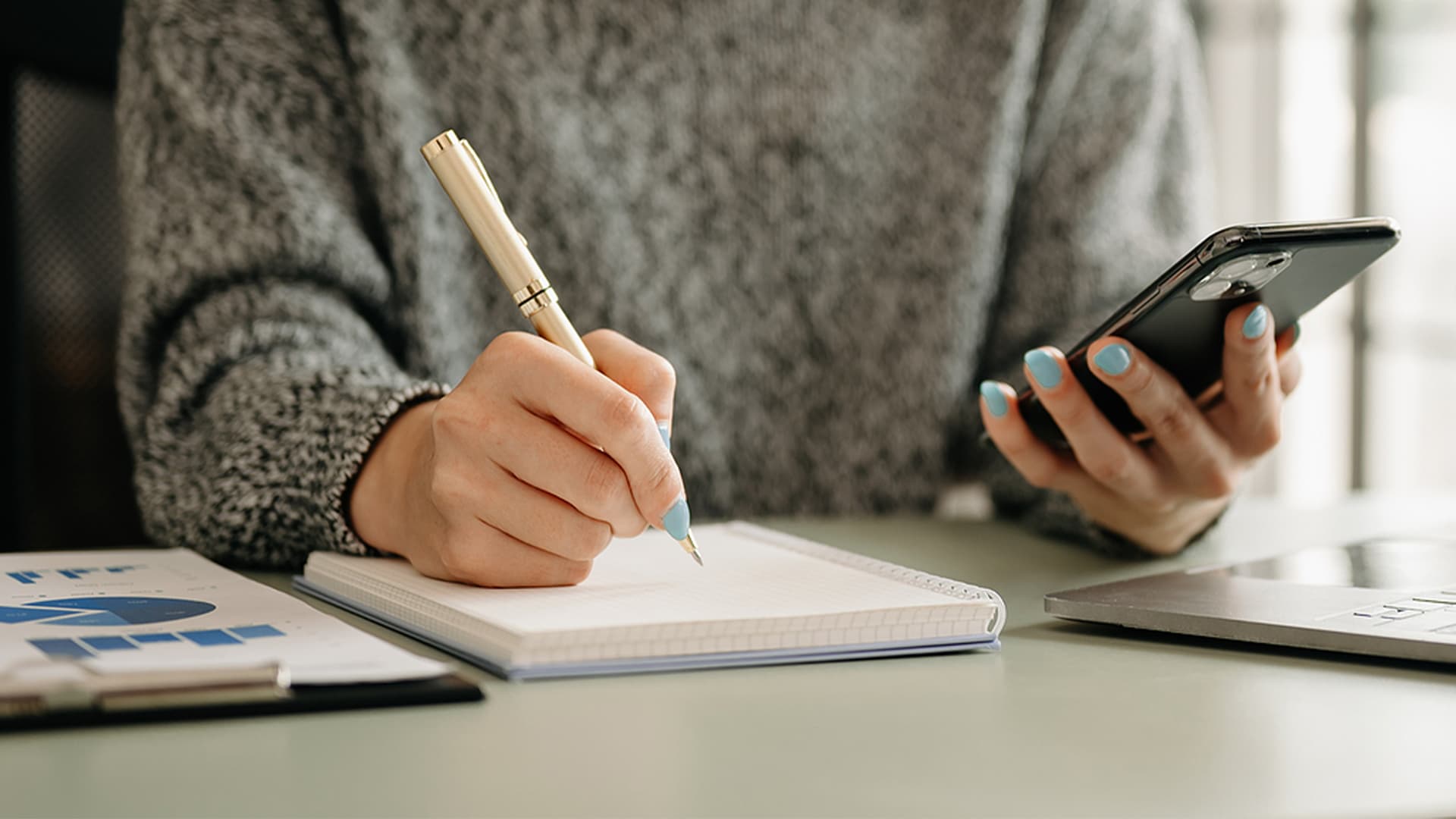 An office worker checks data on a cell phone with a laptop and analytics on the desk
