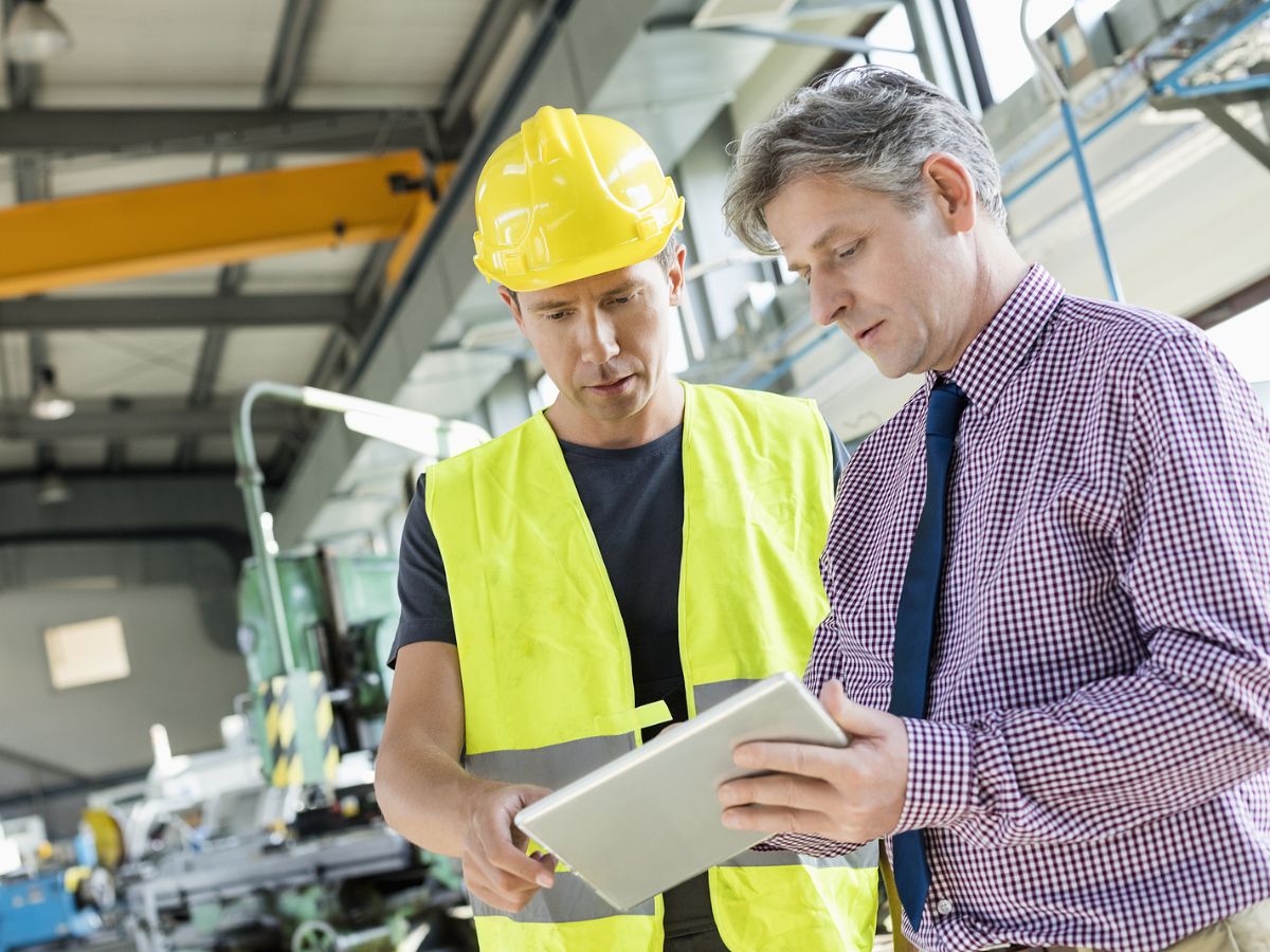 Two Caucasian men look at a tablet on a job site, one in a blue tie and purple checkered dress shirt, the other in a high-visibility vest and hard hat