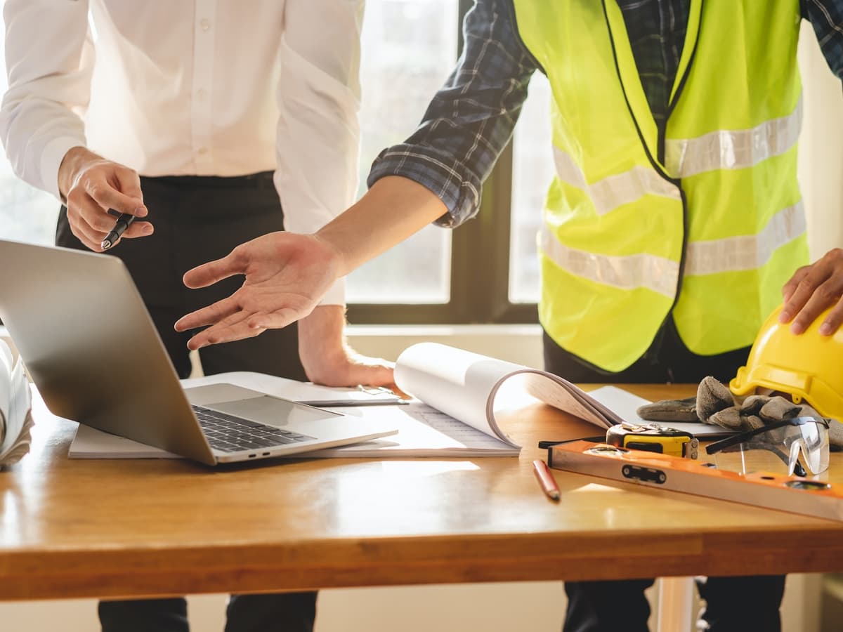 Two individuals in safety vests and hard hats collaborating on a laptop for a construction project.