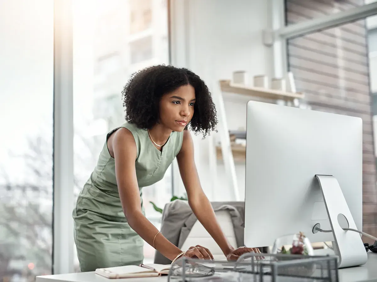 A woman inside of an office and leaning over a desk. She's looking at a computer.