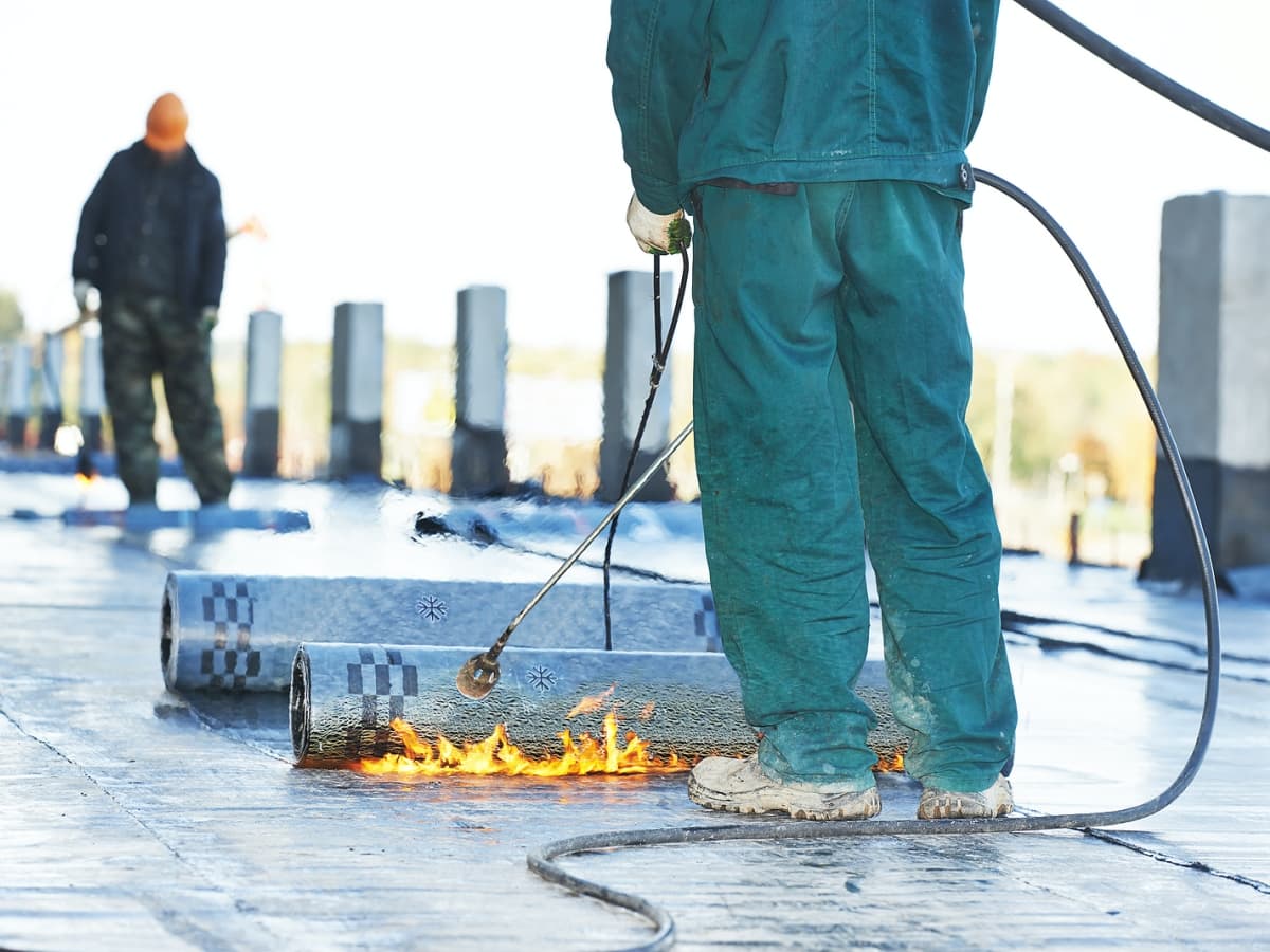 Man in green overalls applying flat roofing with fire tool.