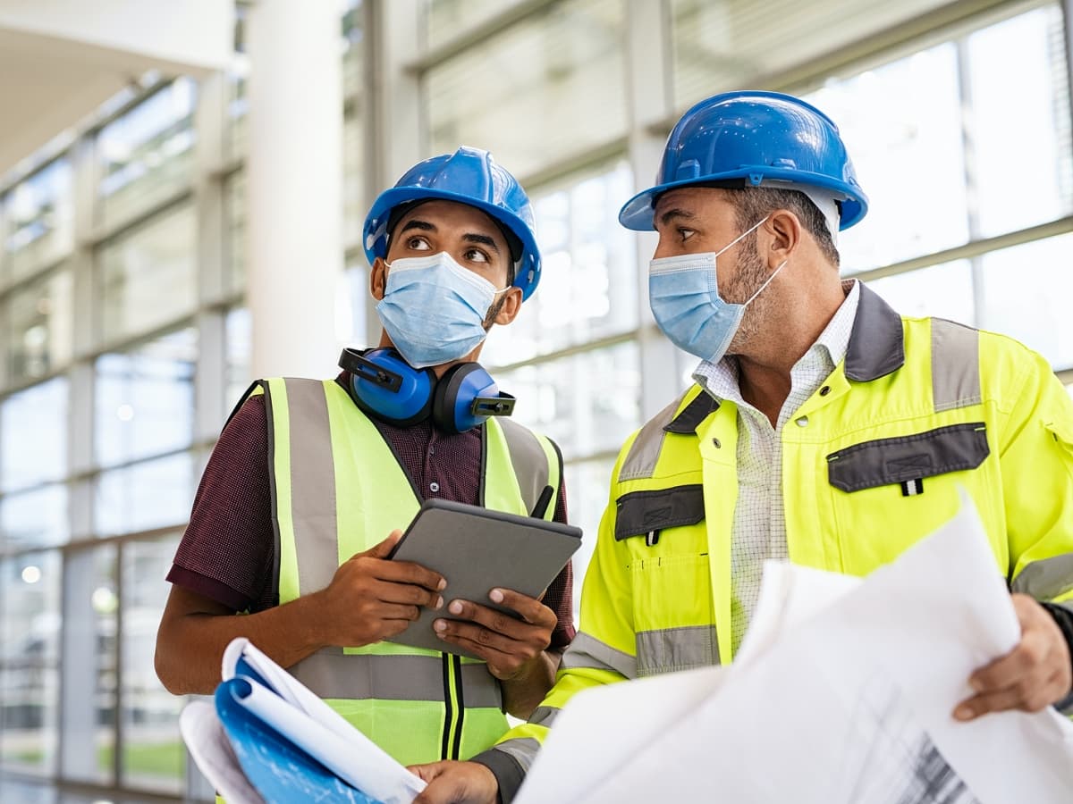 Men in a hard hats and yellow vests focused on laptop while working on a construction site.