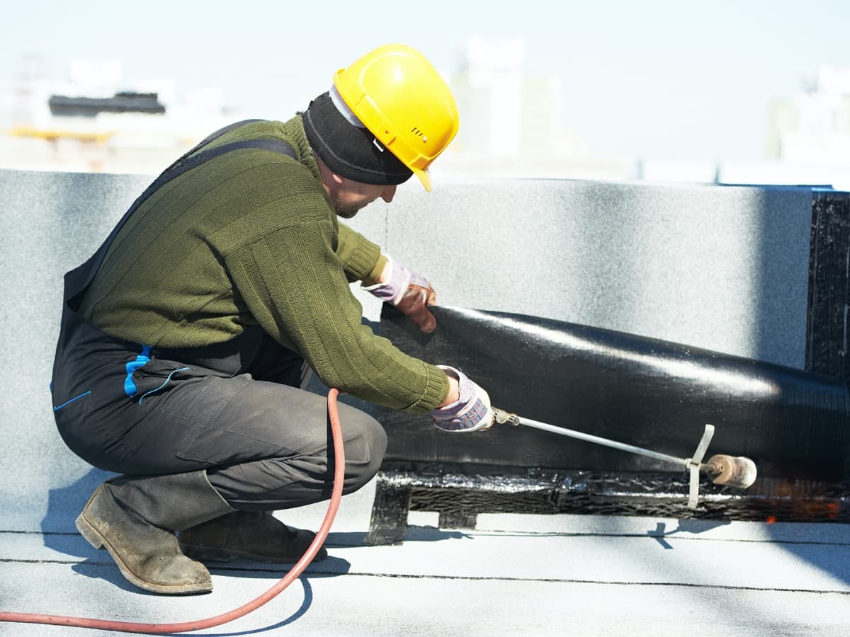 A worker in overalls and a hard hat applies black roofing material.