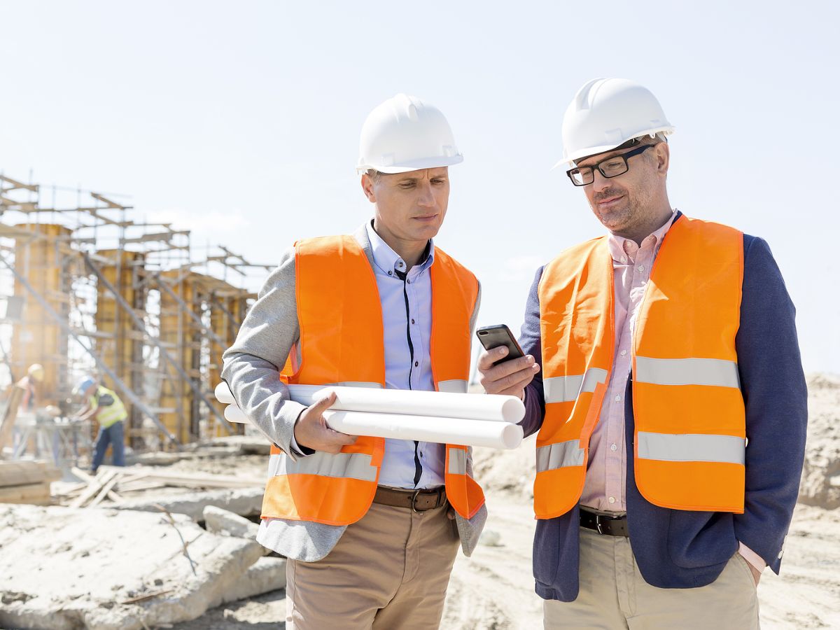Two men on a construction project site stand looking at a mobile phone while wearing high-visibility vests in orange and white