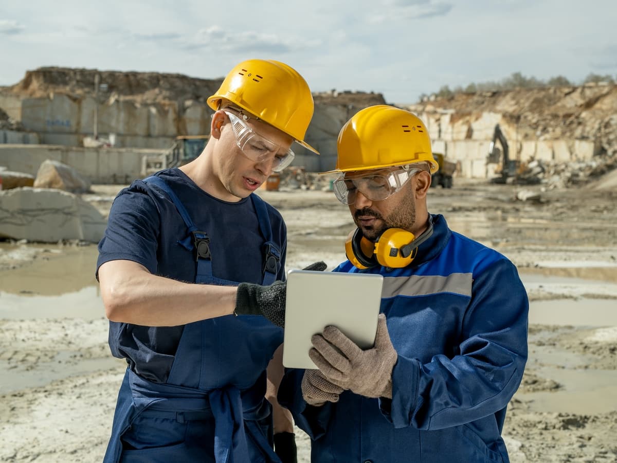 Two construction workers wearing hard hats and safety glasses, focused on a tablet.