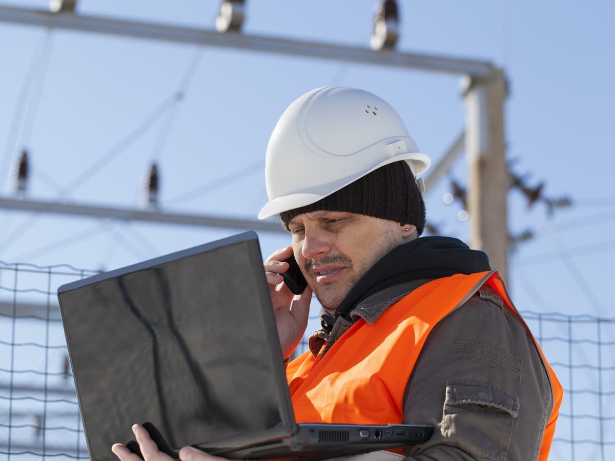 An electrician wearing an orange vest, working on laptop.