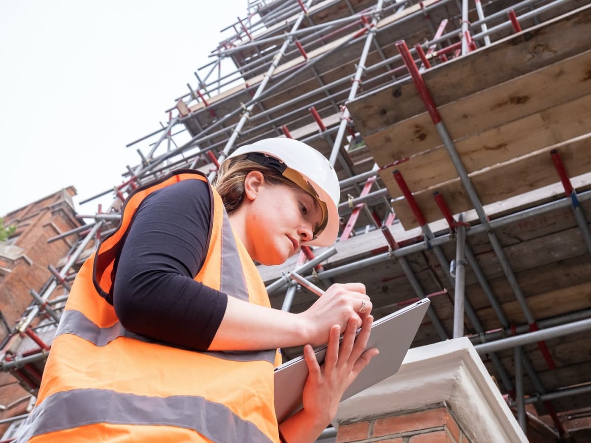 A woman in an orange vest and hard hat diligently writes on a tablet, ensuring accurate documentation.