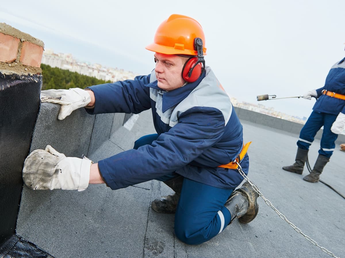 Two contractors on a roof, one holding a tool while the other works with his hands.